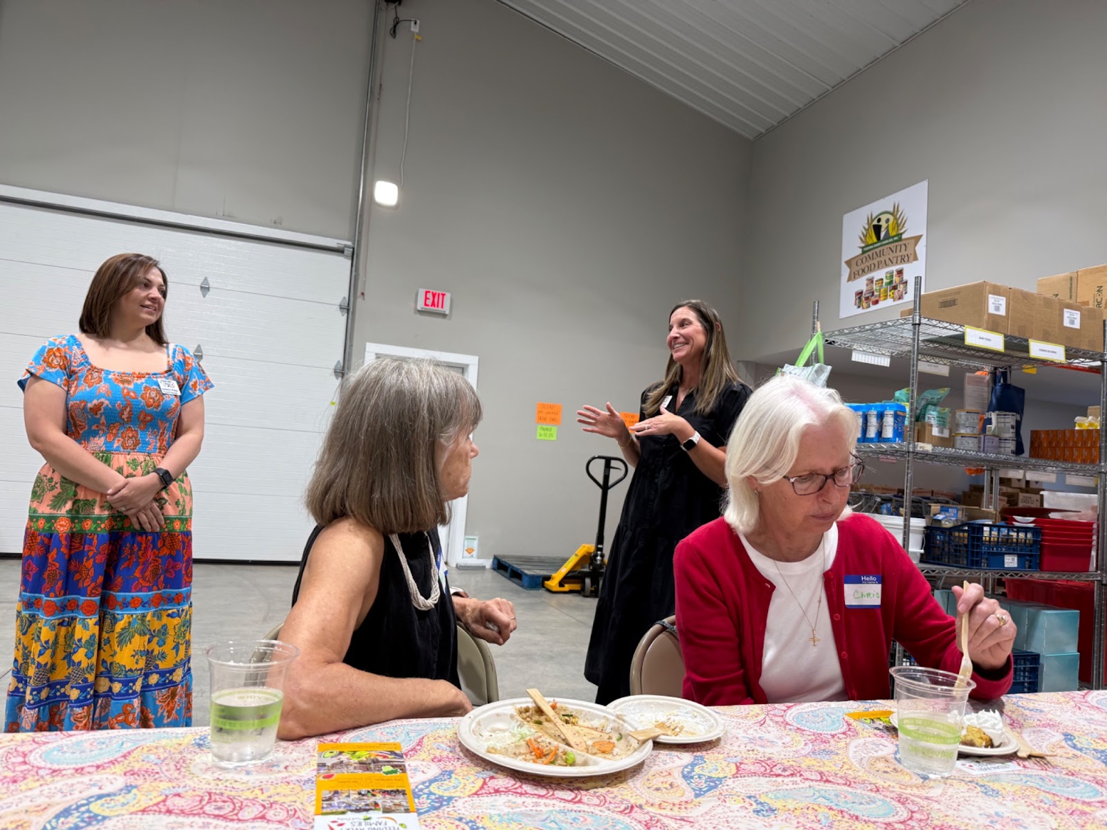 Women gathered around tables in a food pantry warehouse, sharing a potluck meal with volunteers and food distribution shelves visible in the background.