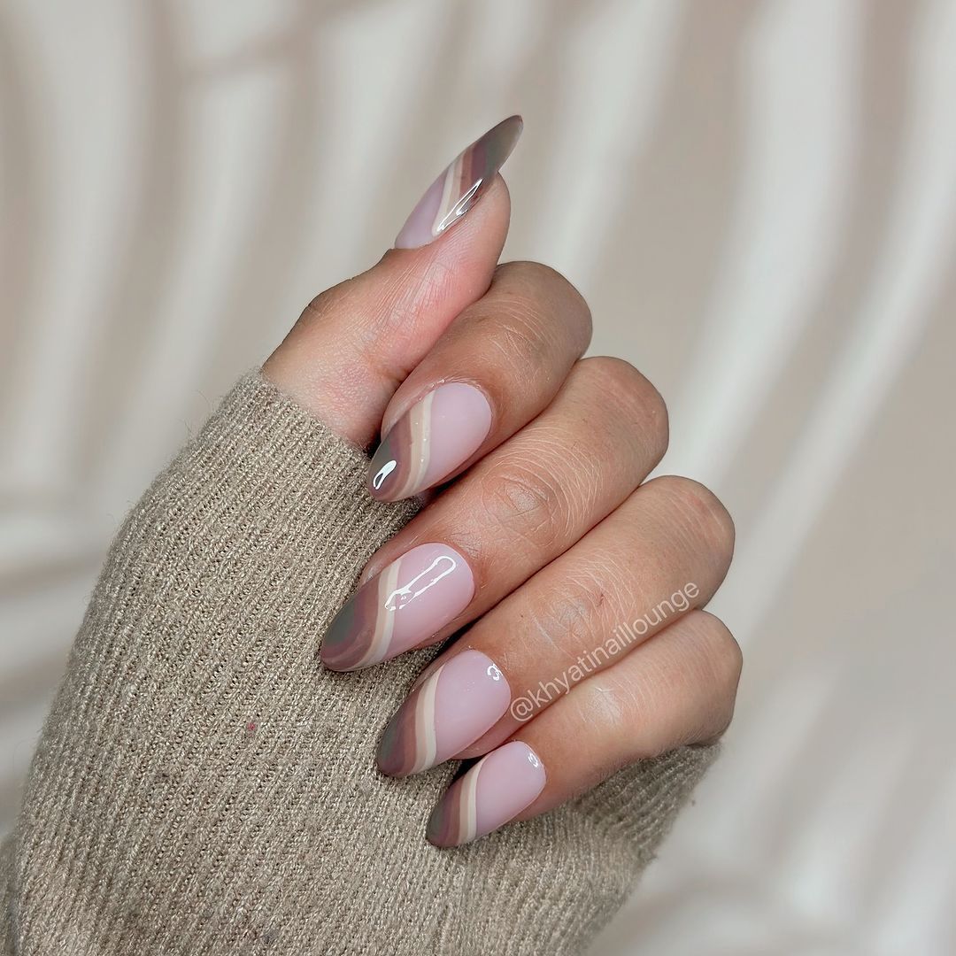 A hand with long, almond-shaped nails featuring a glossy nude and brown geometric nail art design.