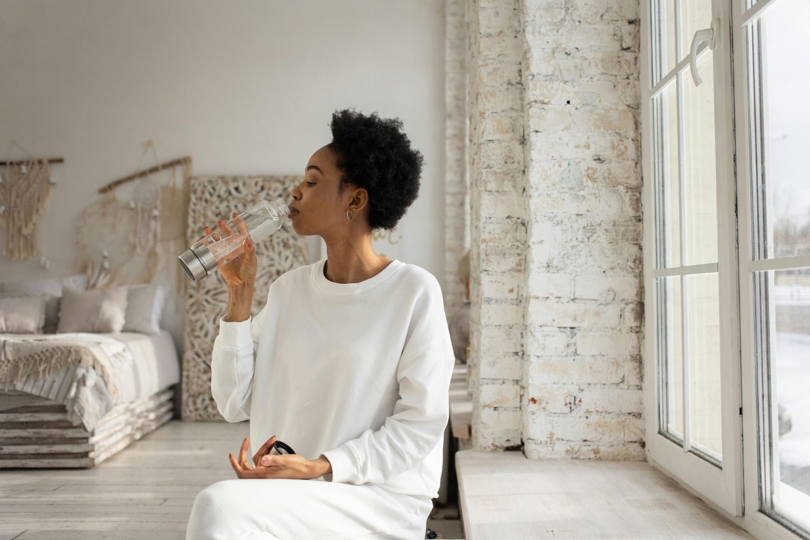 A woman drinks water from a carafe by a window.