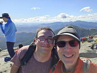 Two men (one young, one older) smiling for the camera on the summit of a mountain, with more mountains visible behind them in the distance.