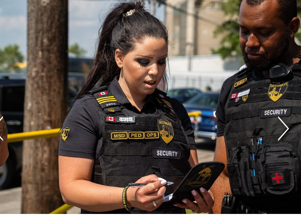 Two security officers stand outdoors; one woman is holding a clipboard and writing while the man beside her looks on. Both wear black uniforms, showing their badges and security labels—reminding others to obtain a security license for such roles.