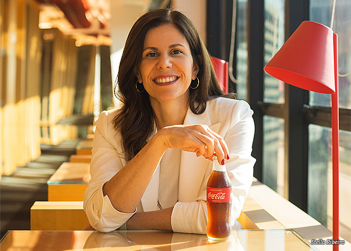 Luciana Starczarini Batista sebtada em um refeitório, segurando uma garrafa de coca-cola gelada e sorrindo para a camera. 
