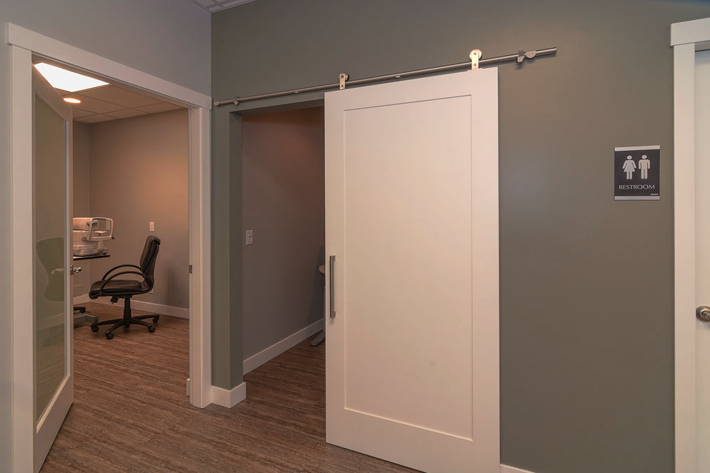 A sliding barn door in a medical clinic with a door to an examination room to the left of it and a sign for the washroom on the wall to the right. 