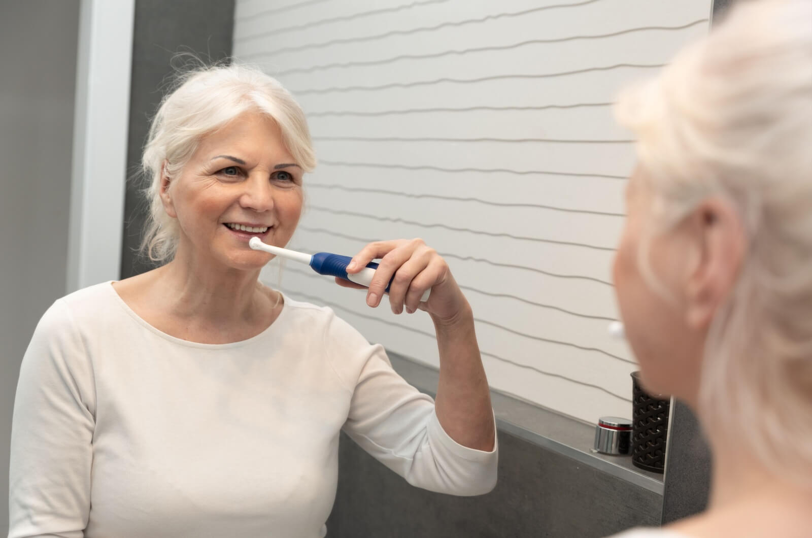 A smiling older adult looking in the mirror with an electric toothbrush in their right hand.