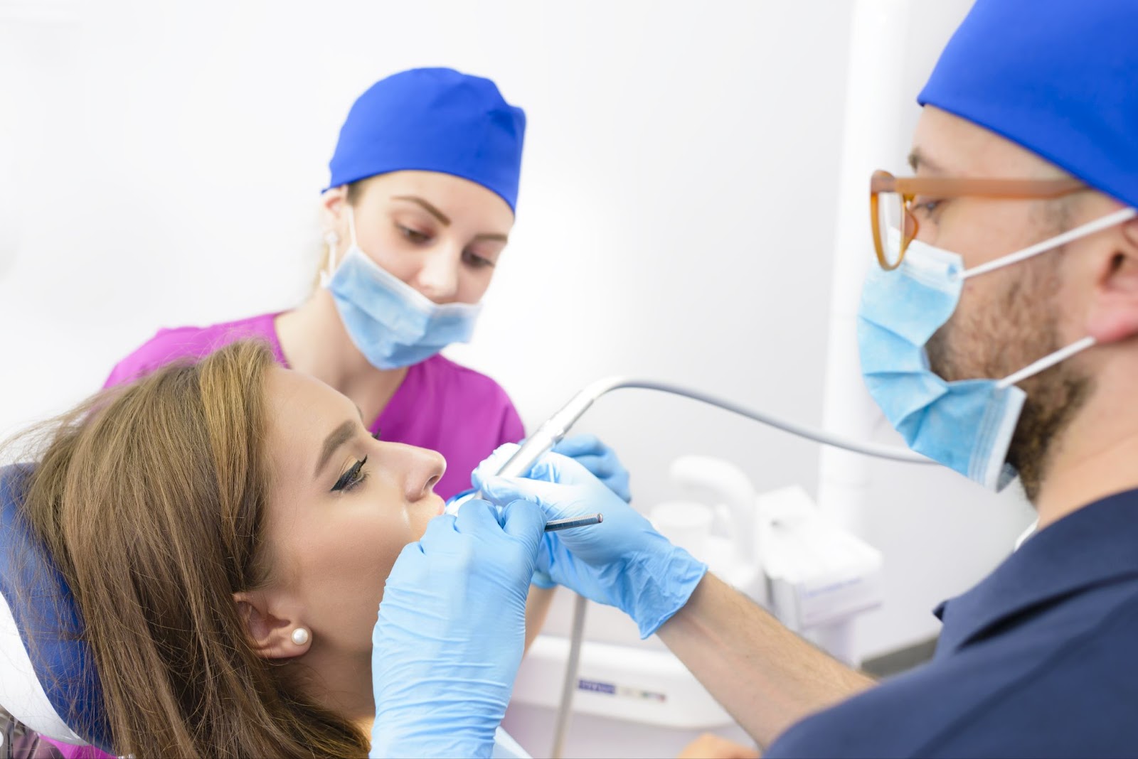 2 dentists lean over a patient, holding dental tools. The person is at ease and relaxed and both dentists are smiling.