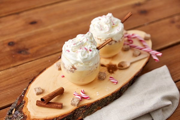 Two mugs of vanilla mousse, garnished with cinnamon sticks and candy canes, served on a rustic wooden board with sugar cubes