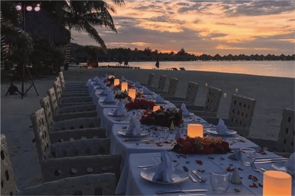 Beachside wedding venue at sunset with long table setup, glowing table lights, and floral accents.
