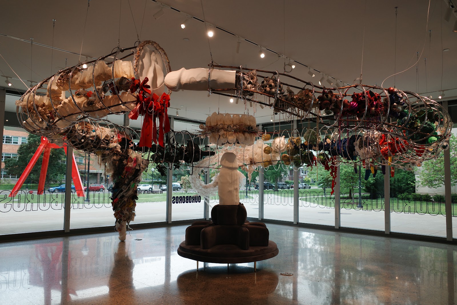 View of Ouroboros exhibition, plastic metal ring sculpture spanning the room hanging from the ceiling, plaster phallus sculpture in center of floor pointing upwards.