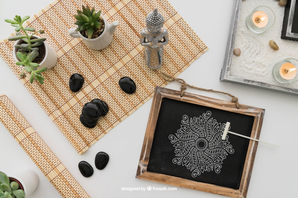 yoga room decor: A flat lay composition with various objects suggesting a theme of relaxation or meditation. There is a small chalkboard with a white mandala design, three lit candles, smooth black stones, two potted plants, and a small Buddha statue. The items are arranged neatly on a light surface with two bamboo mats underneath some of the objects.