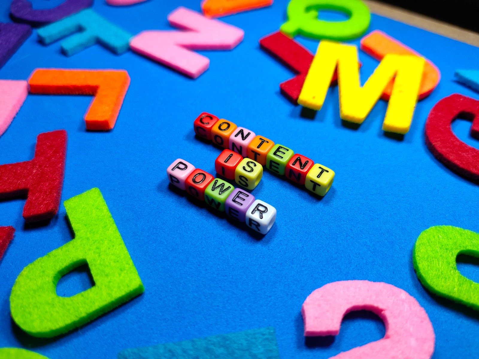 Colored dice spelling out the phrase, “Content is power,” on a blue desk surrounded by other felt letters.