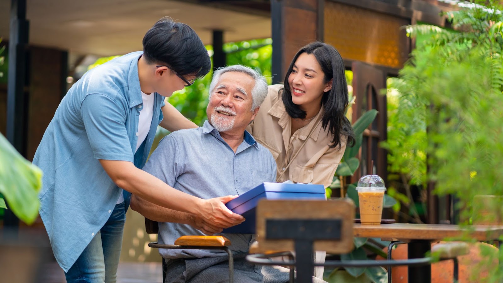 2 young family members visiting an older loved one and smiling as they present him with a gift as he sits at a table outside