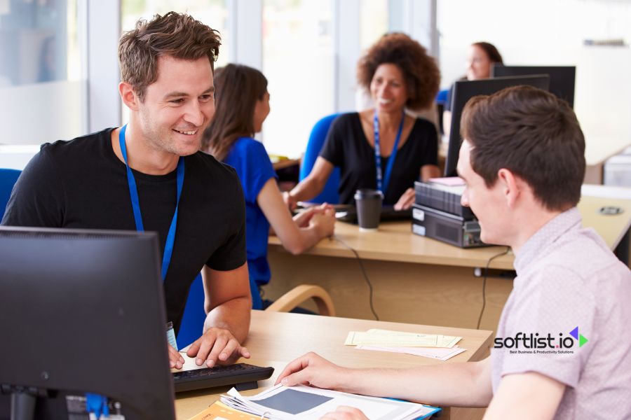 Two men talking and working at a desk in an office.