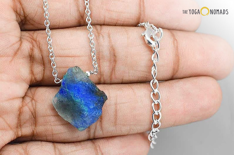 A hand holding a pendant with a silver chain. The pendant features a raw, unpolished stone of deep blue color with some lighter blue and greenish hues, which could be identified as Azurite. The stone is irregular in shape and has a natural, rough texture.