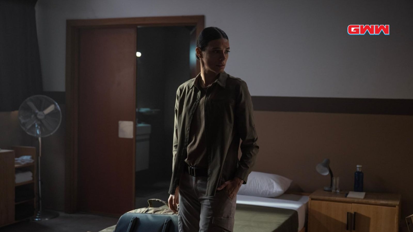 Laysla De Oliveira portraying Cruz, stands inside a dimly lit room looking serious.