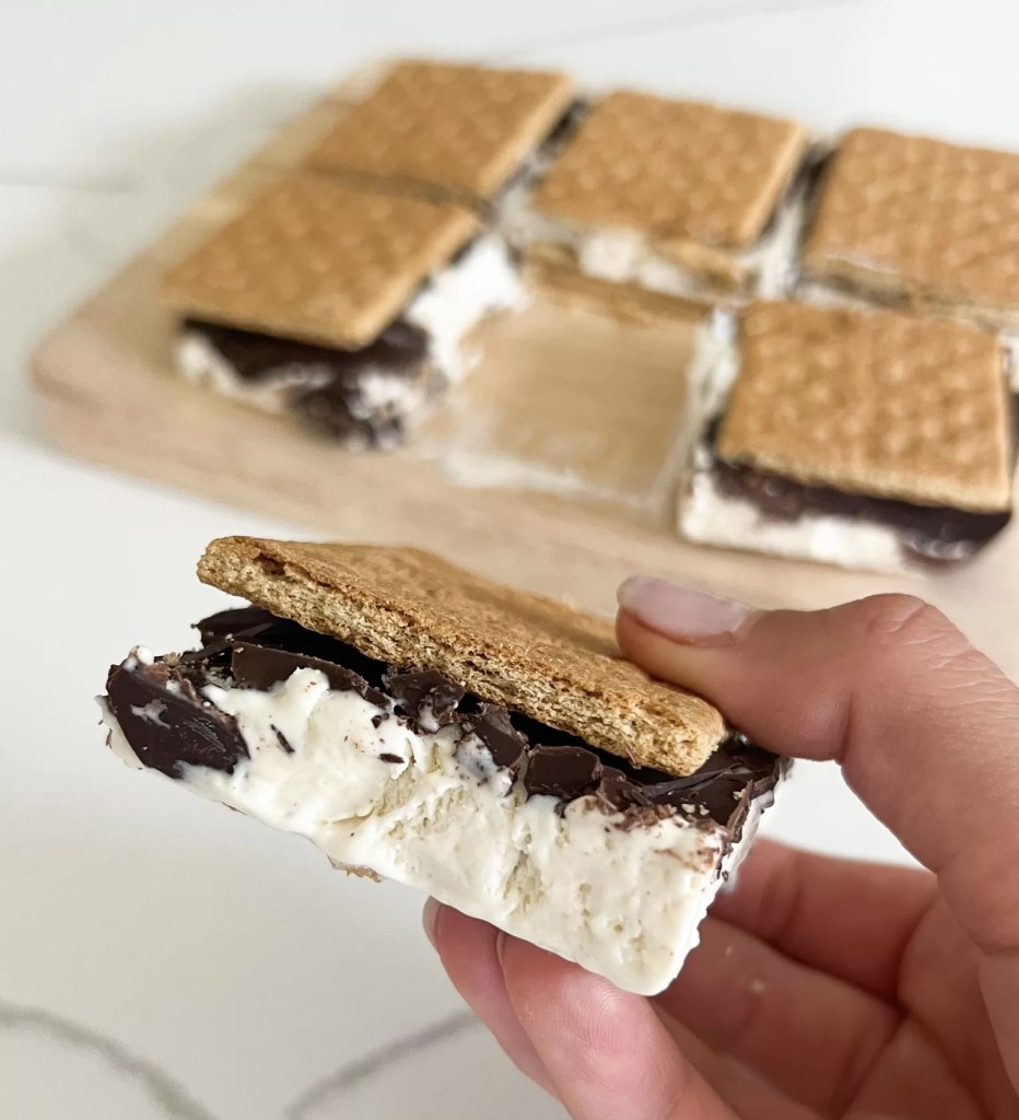 These frozen protein s'mores bars are gluten free, dairy free, high protein, can be low sugar, the perfect summer dessert. On a cutting board are the s'mores bars before cutting. Three golden brown graham crackers with hardened rich chocolate showing through the cracks between on the sides. A creamy, thick white protein whip shows spilling over the sides. Like ice cream sandwiches and ready to eat. One is cut into a square and showing side angle of the thick whip layer, chocolate crunch, and cracker 