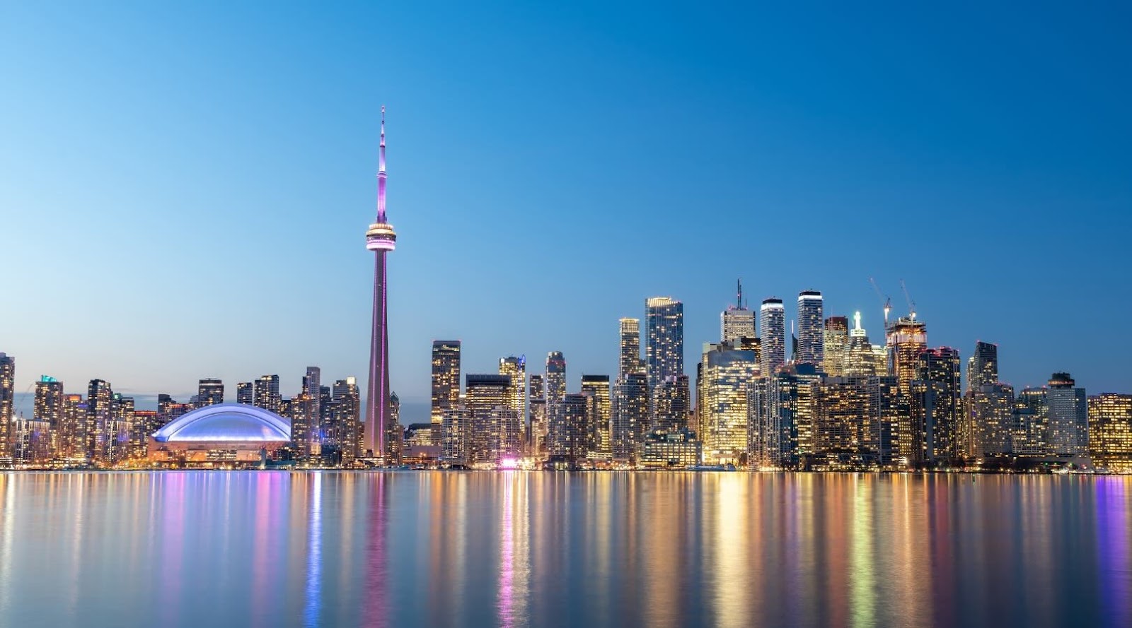 La ligne d'horizon de Toronto au crépuscule, avec la Tour CN illuminée en violet et les lumières de la ville se reflétant sur les eaux calmes du lac Ontario. 