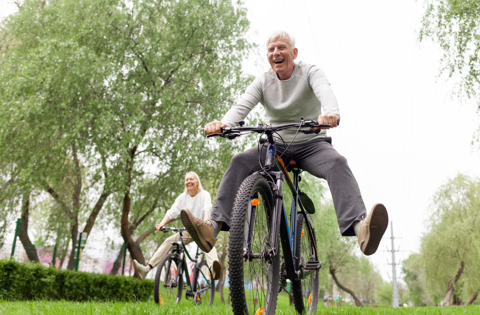 2 seniors goofily ride their bikes in a local park, kicking their legs out.