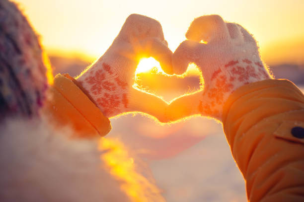 A woman standing outdoors in a snowy winter landscape, making heart-shaped hands.