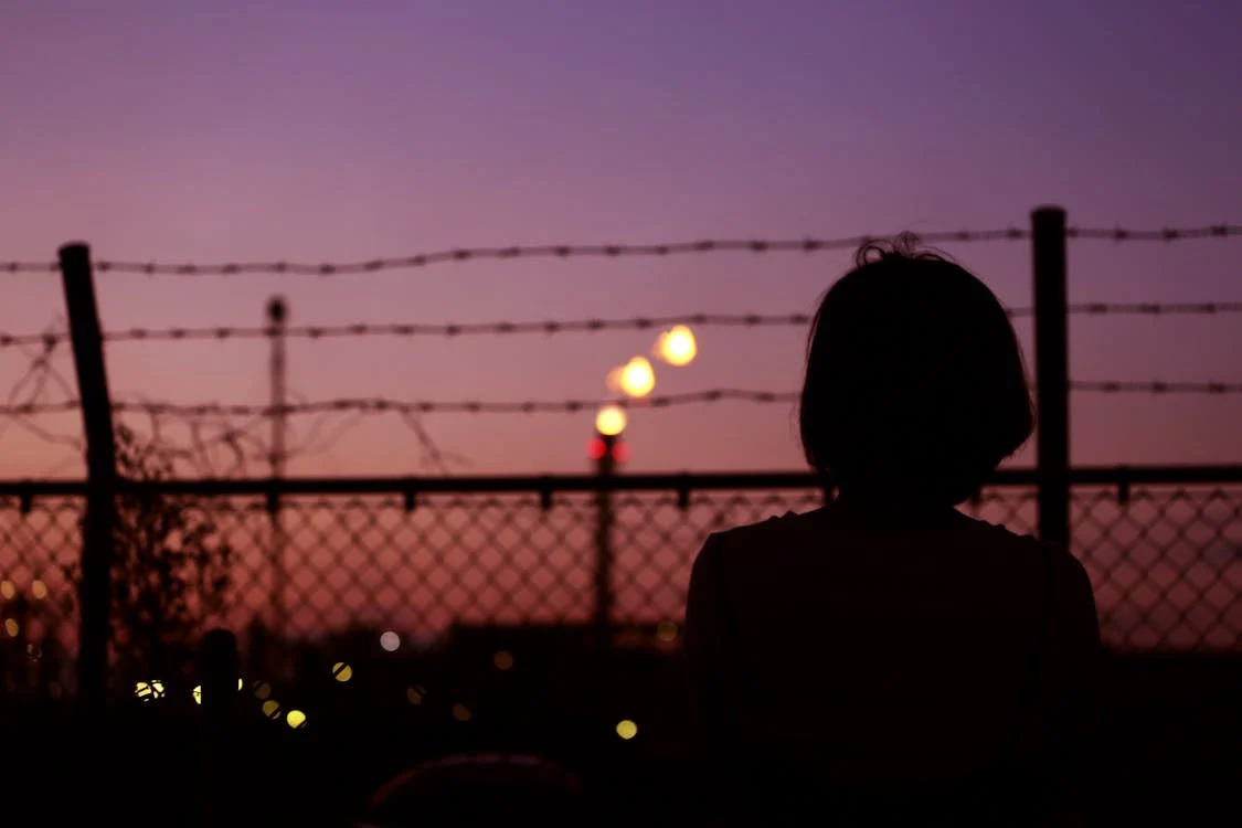 A female figure silhouette against a purple and red sky at sunset. There is a tall mesh fence with barbed wire at the top in front of the woman, as she looks out beyond it.
