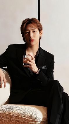 This contains an image of GOT7's BamBam in a suit sitting on a chair holding a drink