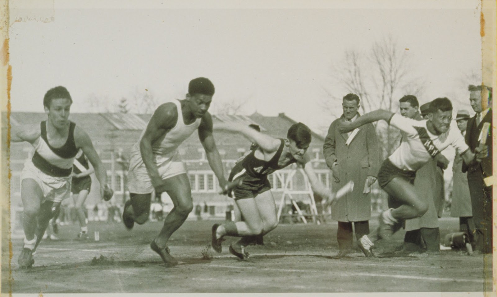 A track race with 4 competitors leaning forward as they run, with several spectators looking on