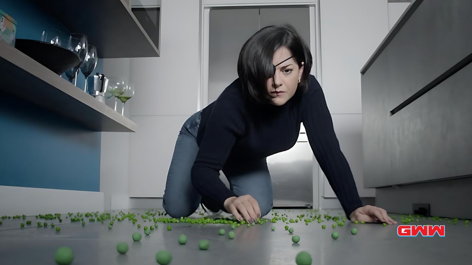 Bibi Garvey crawling on the floor, collecting scattered peas in a kitchen.