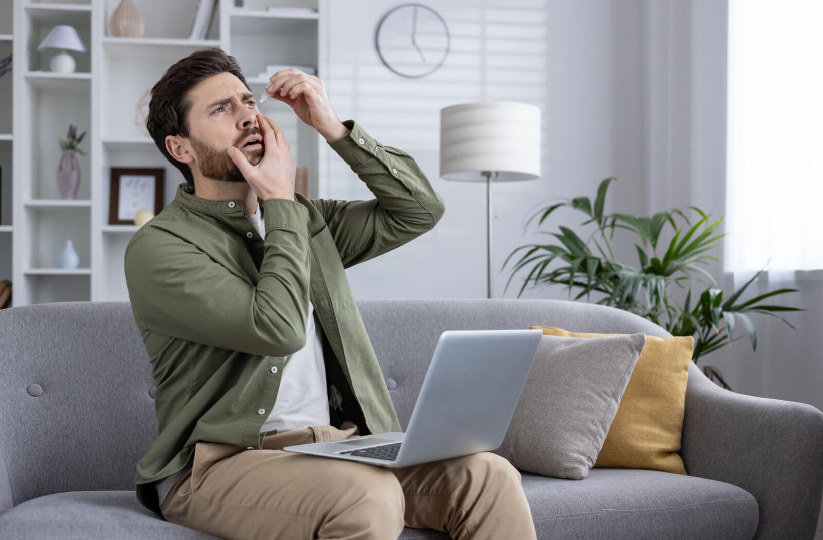 A young professional on their couch at home using eye drops to prevent wintertime dry eye discomfort.