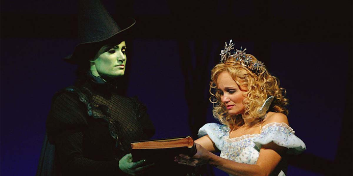 Escena del musical con dos personajes, uno con piel verde sosteniendo un libro y otro con vestido blanco brillante y corona.