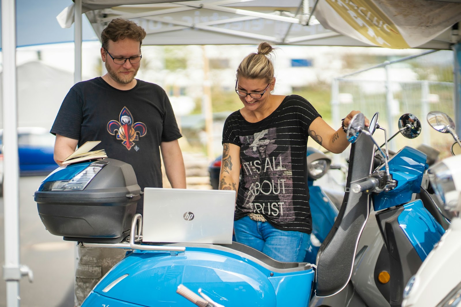 A man and a woman are standing next to a blue scooter under a canopy. Discussing something while looking at a laptop placed on the scooter's seat.