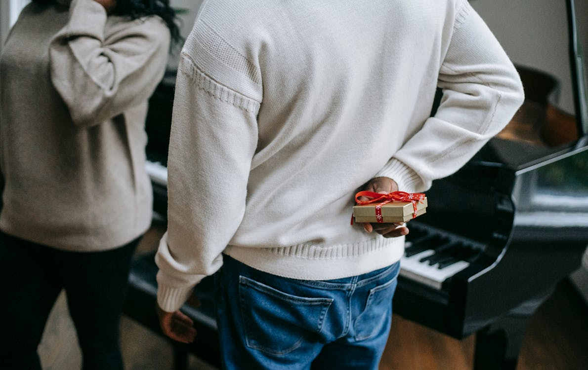 Man in a white sweater hiding a small wrapped Christmas gift behind his back, standing near a woman and a grand piano indoors.