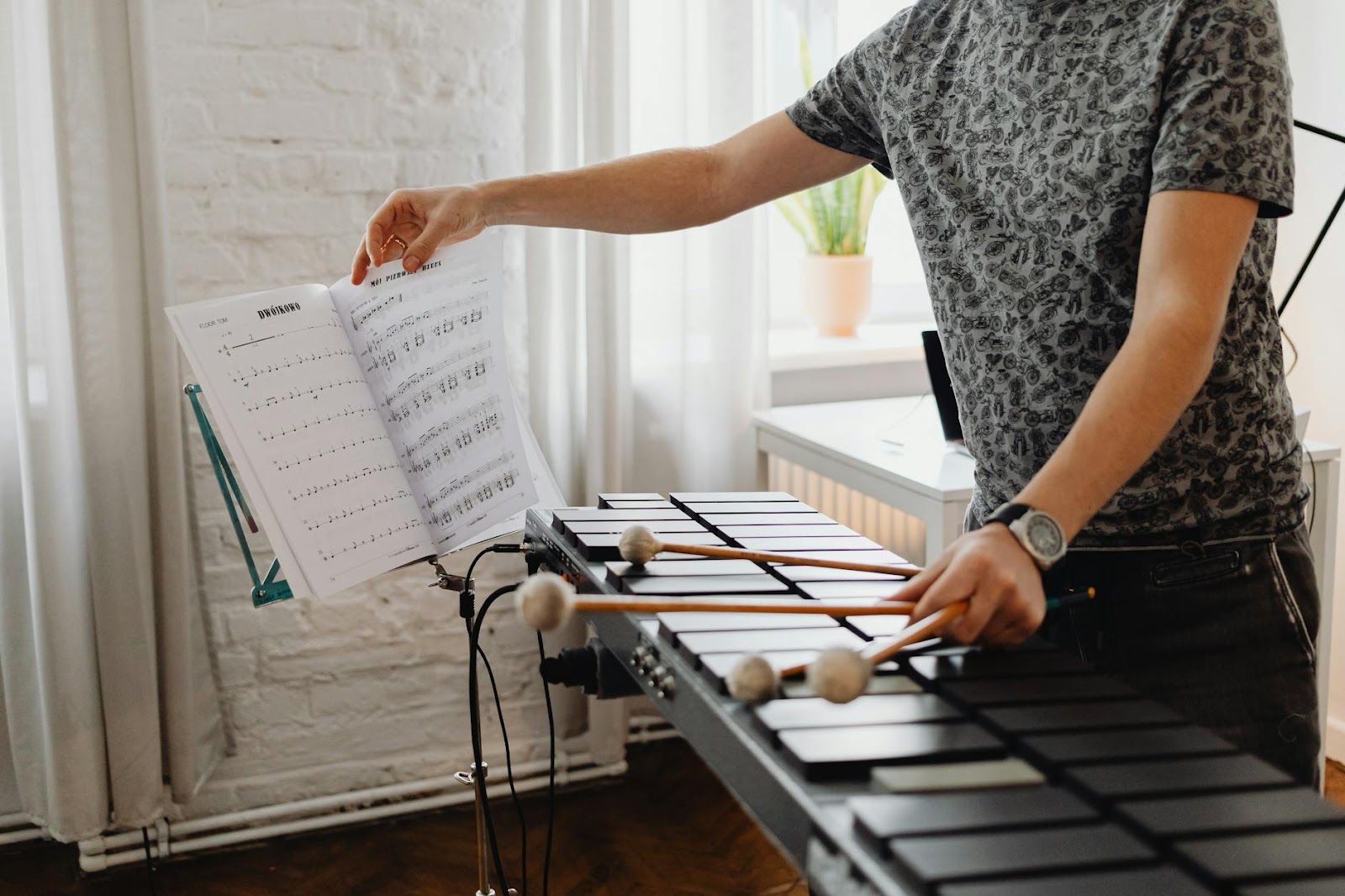 Adult musician with gray shirt playing xylophone indoors, holding mallets and sheet music.