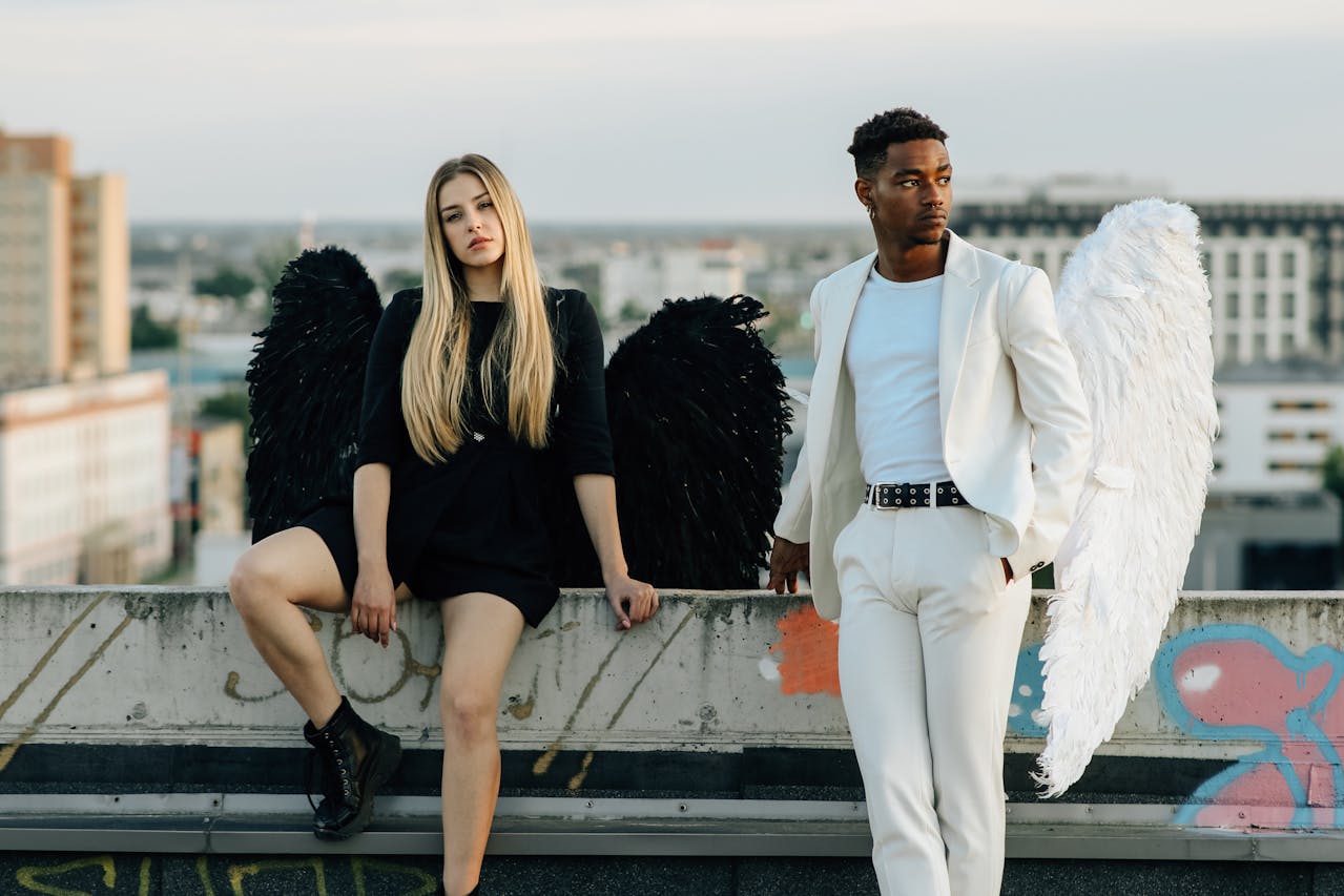 Two individuals with angel wings in a white and black relax on a ledge, enjoying a serene moment together in a dreamy setting.