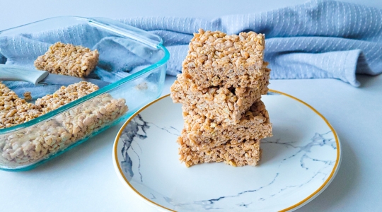 Une pile de carrés de Rice Krispies protéinés sur une assiette à motif marbré, avec un plat de cuisson en verre contenant d'autres carrés en arrière-plan.