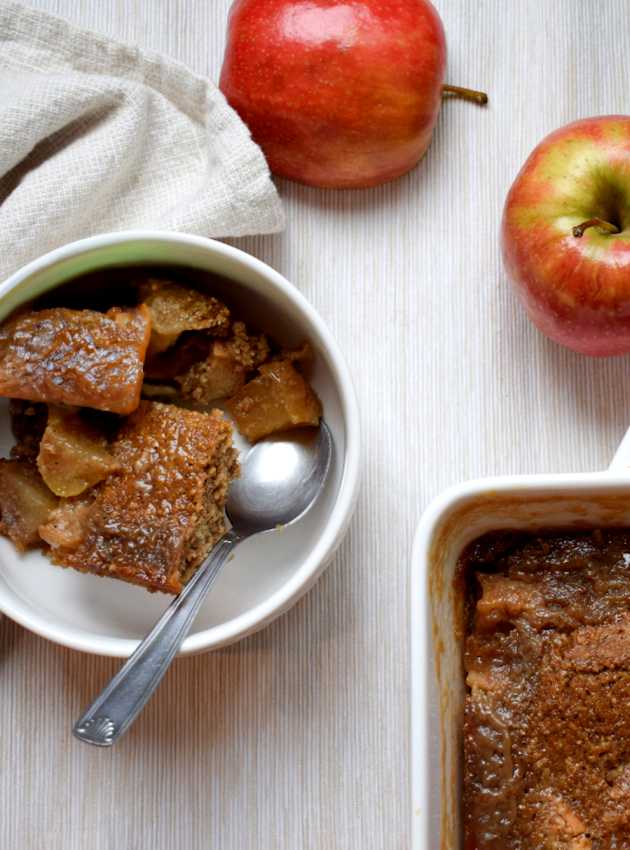 Apple pudding served in a white bowl with a spoon, next to a baking dish and fresh apples.