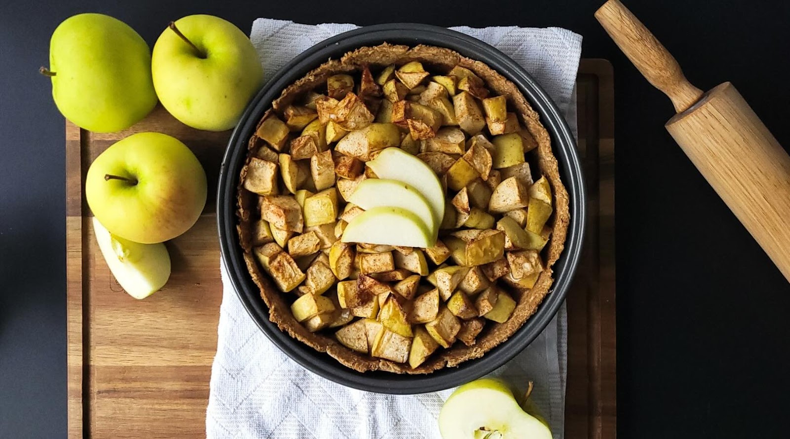 Tarte aux pommes réduite en sucre créé par une nutritionniste-diététiste sur une planche en bois et un linge à vaisselle avec des pommes et un rouleau à pâte