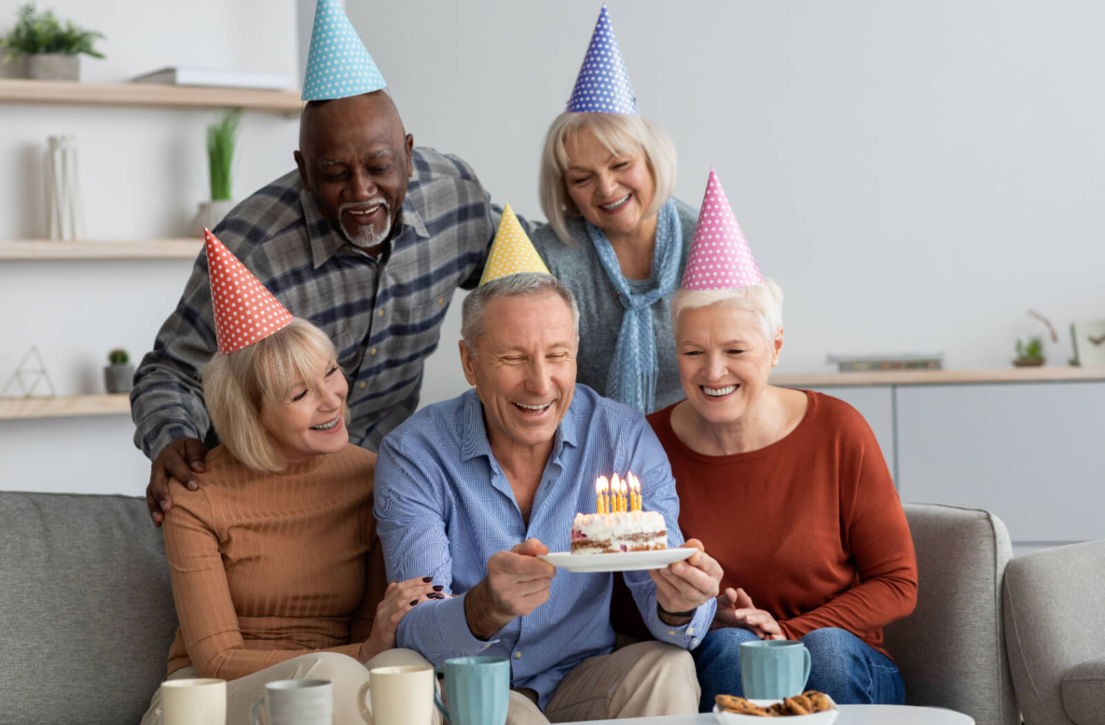 A group of older adults wearing party hats, celebrating a birthday and holding a cake provided by their assisted living community.