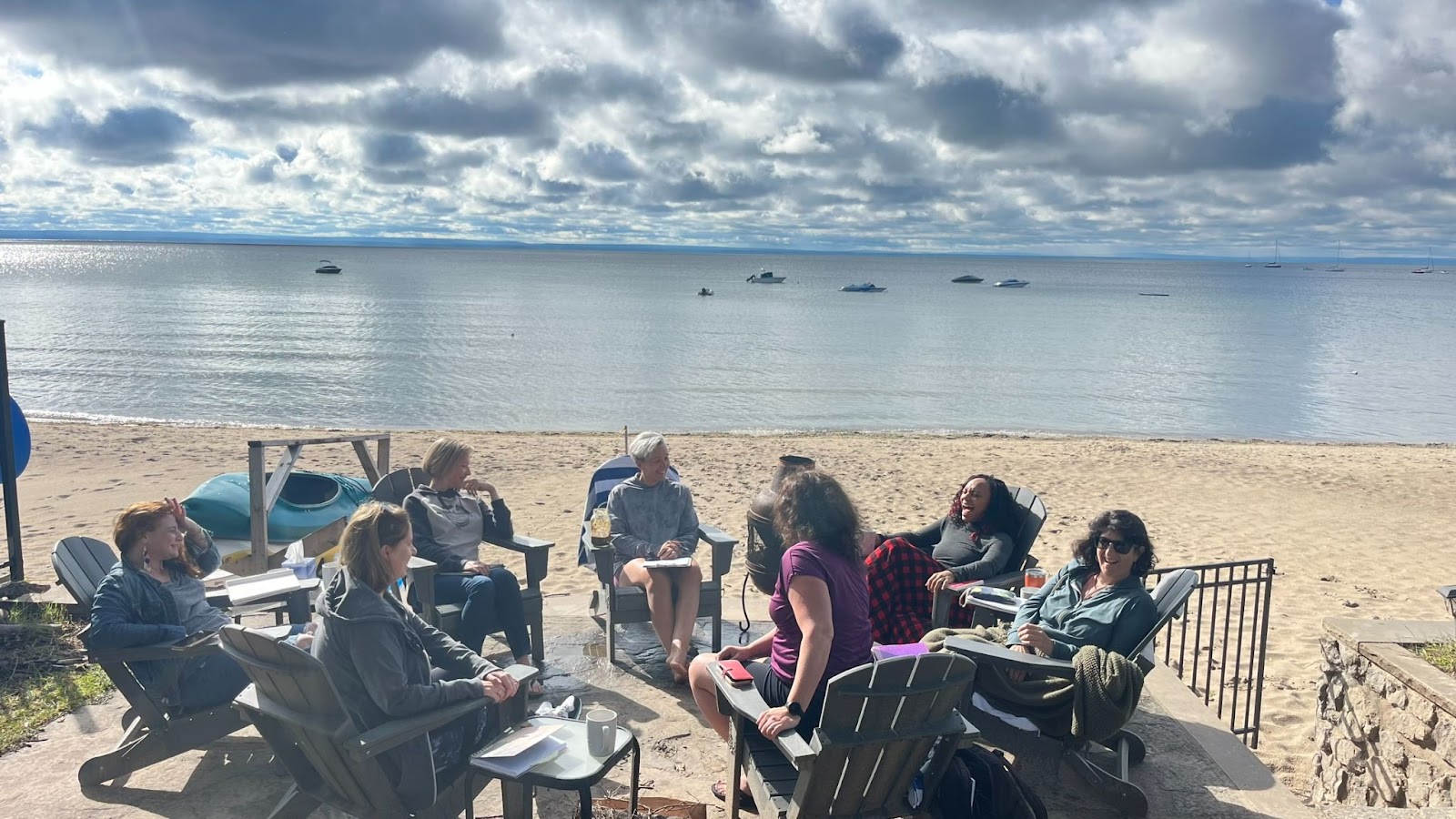 Tarzan and 6 other women sit in Adirondack chairs on a sandy beach, with beautiful Lake Erie in the backdrop, spackled with small boats on calm, shallow water