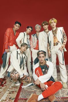 This contains an image of NCT
group members sitting next to each other on top of a rug in front of a red wall