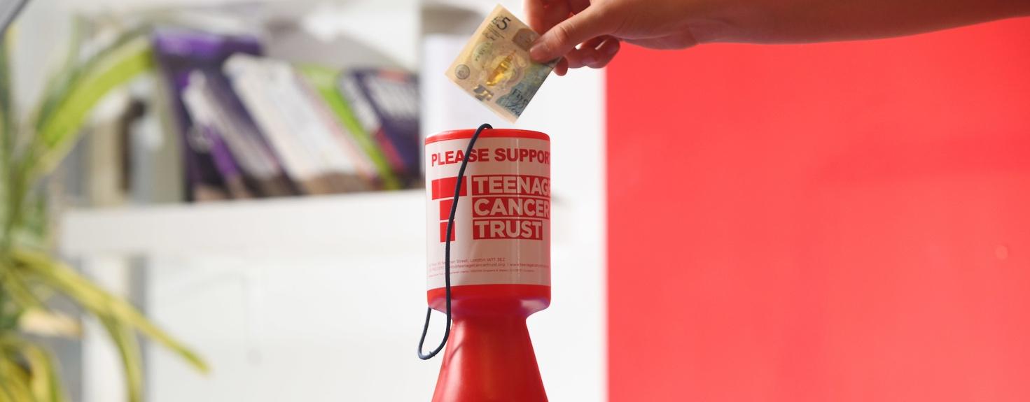 A hand putting money into a red container Description automatically generated