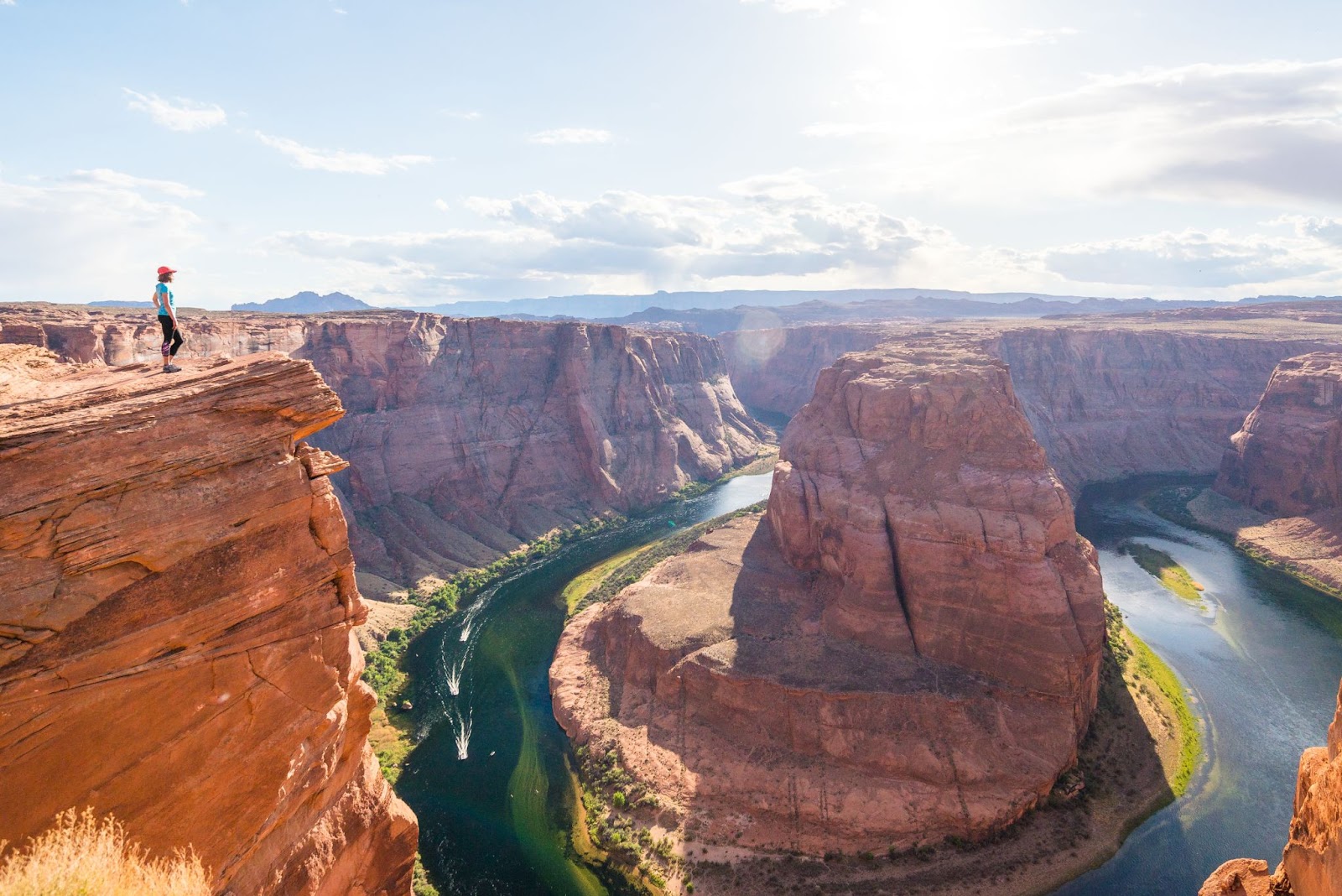 A girl in an orange hat stands at a ledge on the far left overlooking Horseshoe Bend, part of the Grand Canyon, on a sunny day