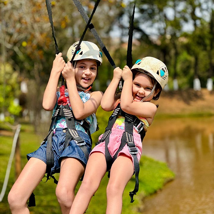 Duas meninas se divertindo em um passeio de tirolesa ao ar livre, usando capacetes e cintos de segurança, com árvores e natureza ao fundo.