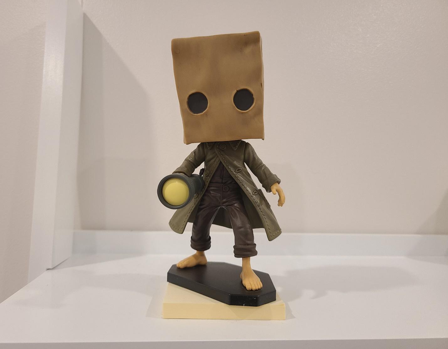 A plastic figurine of a guy wearing a brown paper bag over his head, with two black circular eyes peeping out. He is wearing a trench coat and wielding a flashlight, photographed on a white shelf.