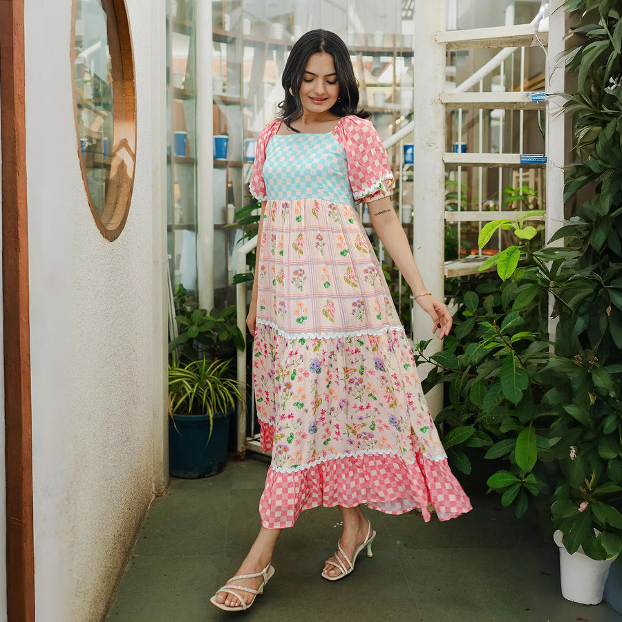  Woman wearing a pastel patchwork maxi dress with floral and checkered prints, posing gracefully in a garden-like indoor setting.