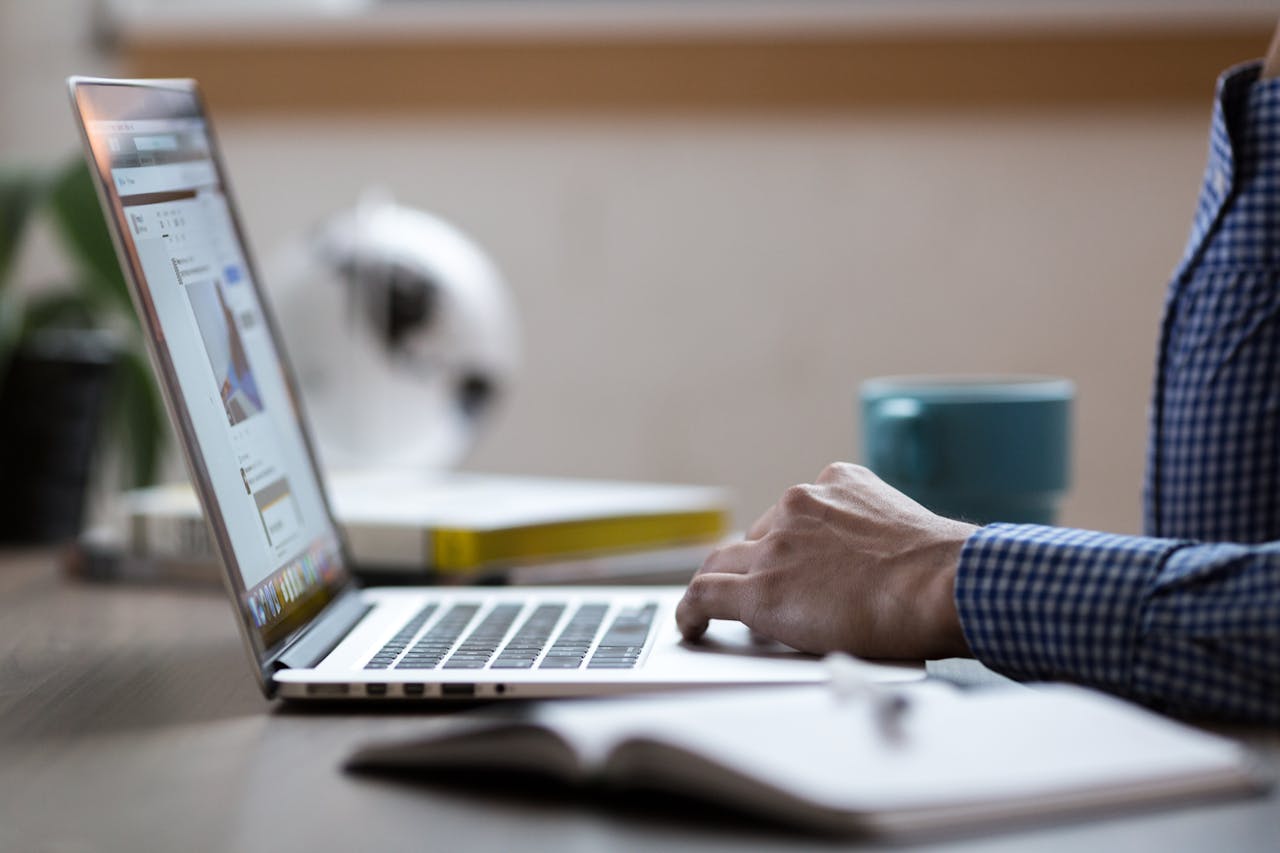 Close-up view of a person working on a laptop, with a notebook and a coffee mug on the desk, in a modern workspace setting.