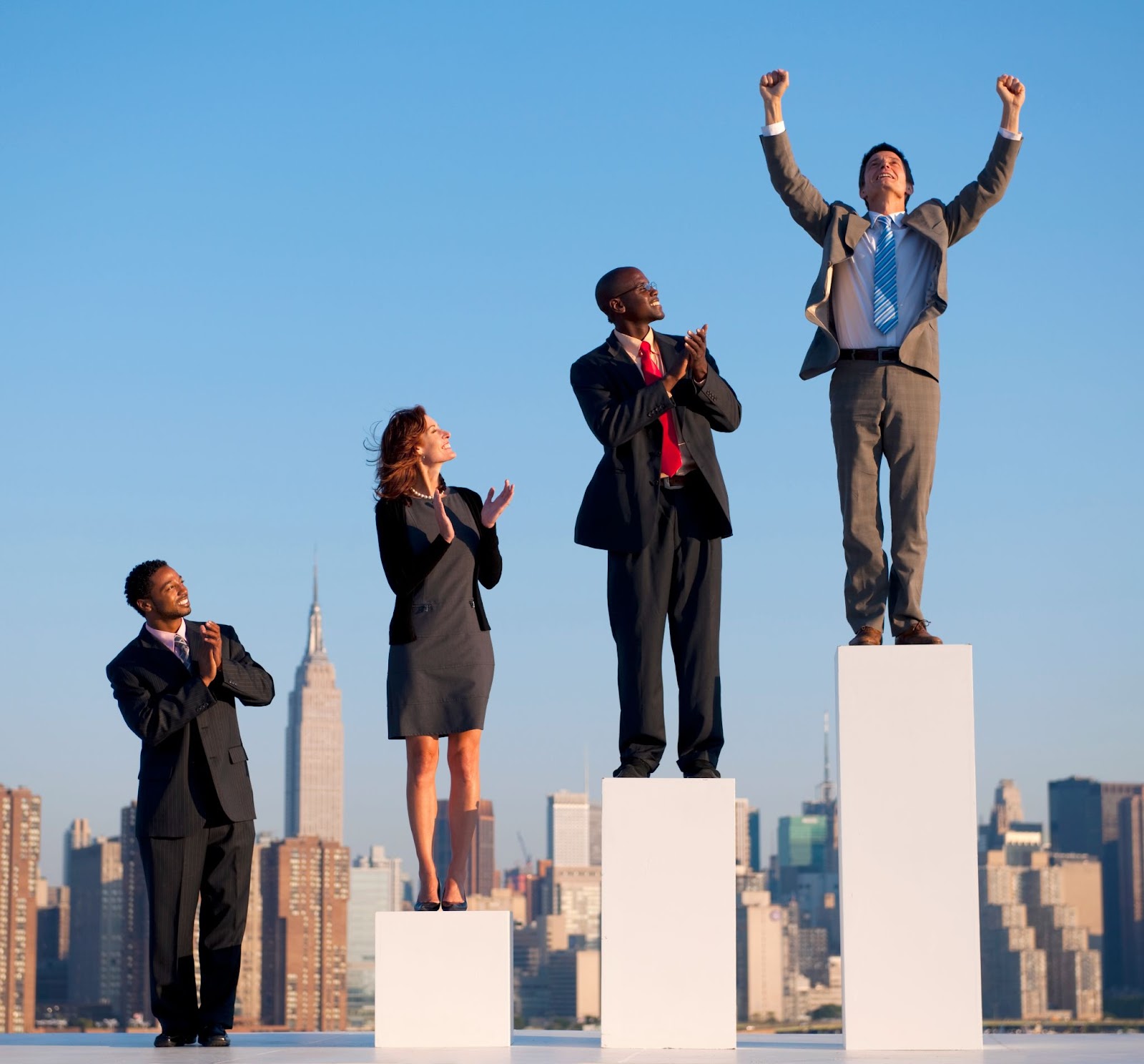 Group of professionals in business attire celebrating a colleague’s success. One man stands triumphantly on a podium with raised arms, while the others stand below, clapping and smiling with a city skyline in the background.