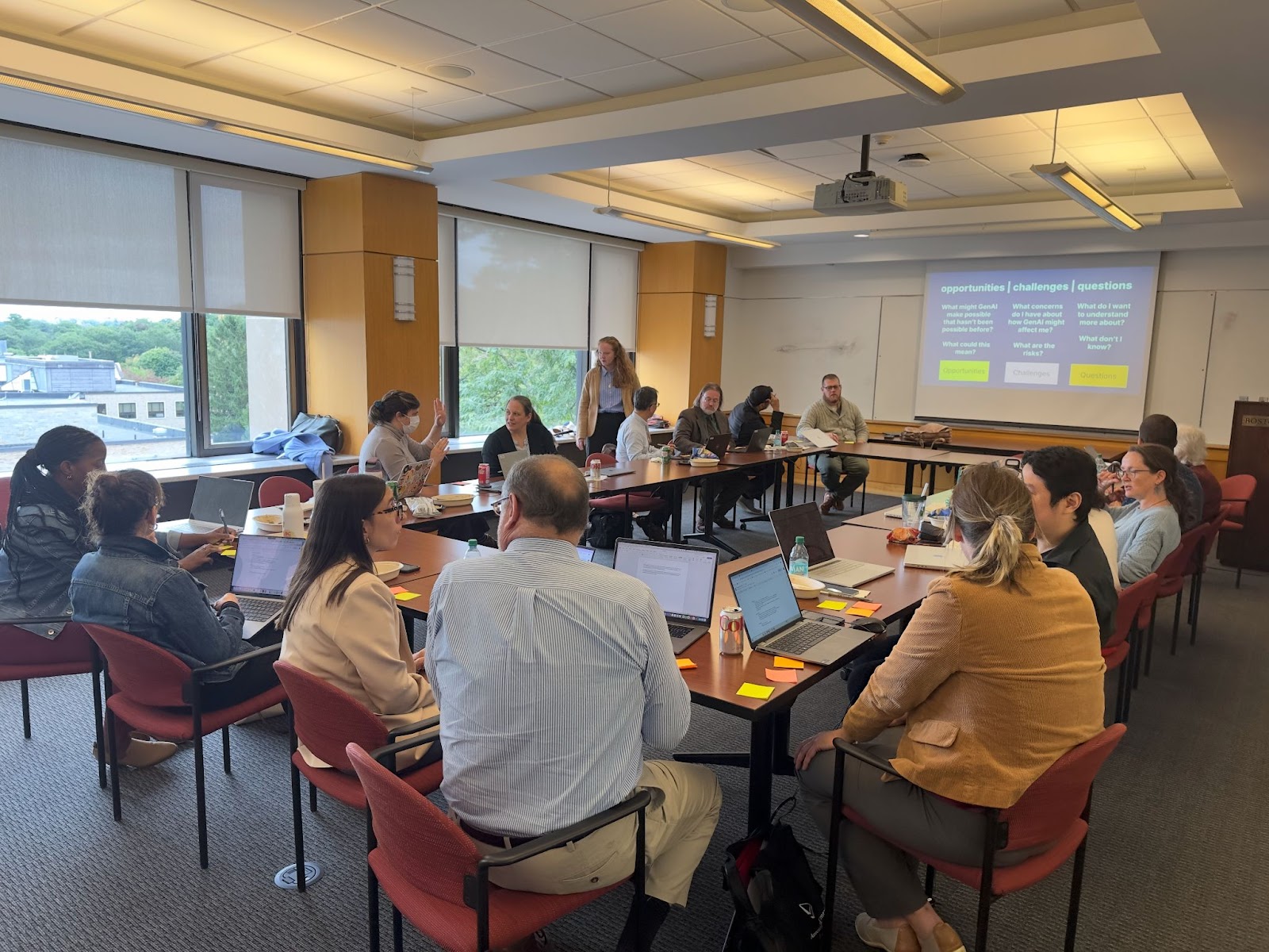 A group of people are seated around tables in a conference room, engaged in a discussion with a presentation displayed on a screen.