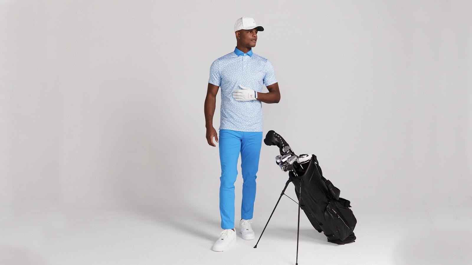 Man wearing a blue golf attire is stand with a golf bag leaning against him with a grey backdrop.