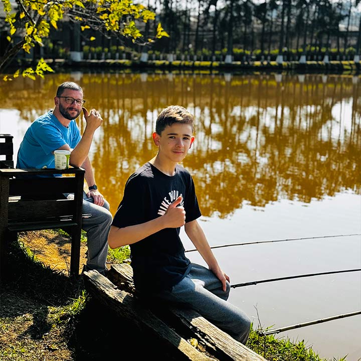 Homem e menino pescando juntos à beira de um lago, aproveitando um momento de lazer ao ar livre, com árvores ao fundo e uma tarde ensolarada.