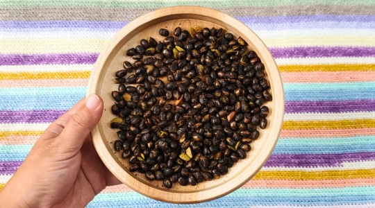 Un bol en bois rempli de haricots noirs rôtis, posé sur un tapis rayé coloré.