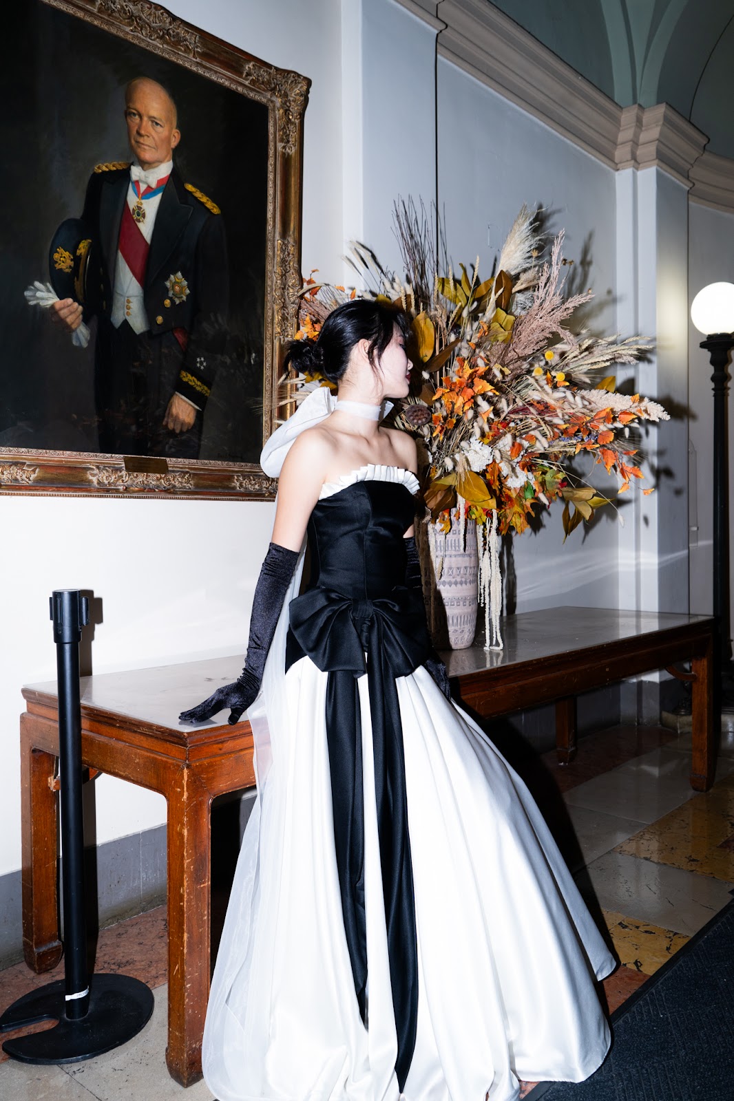 Enkhjin Batbold poses in her black-and-white three-piece gown, standing beneath a portrait and surrounded by autumn floral arrangements. 